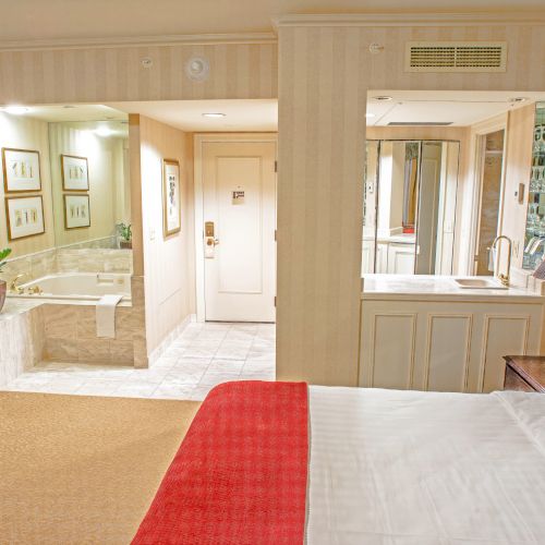 A hotel room with a bed, red throw, beige walls, a seating area with a red chair, plant accents, and an open bathroom/closet area connected to a bright hallway.