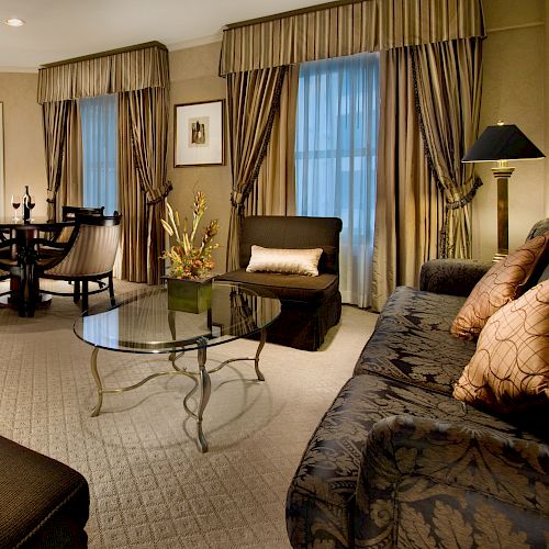 A luxurious hotel suite with a seating area, sofa, armchairs, glass coffee table, round dining table, chandeliers, curtains, a TV, and warm lighting.