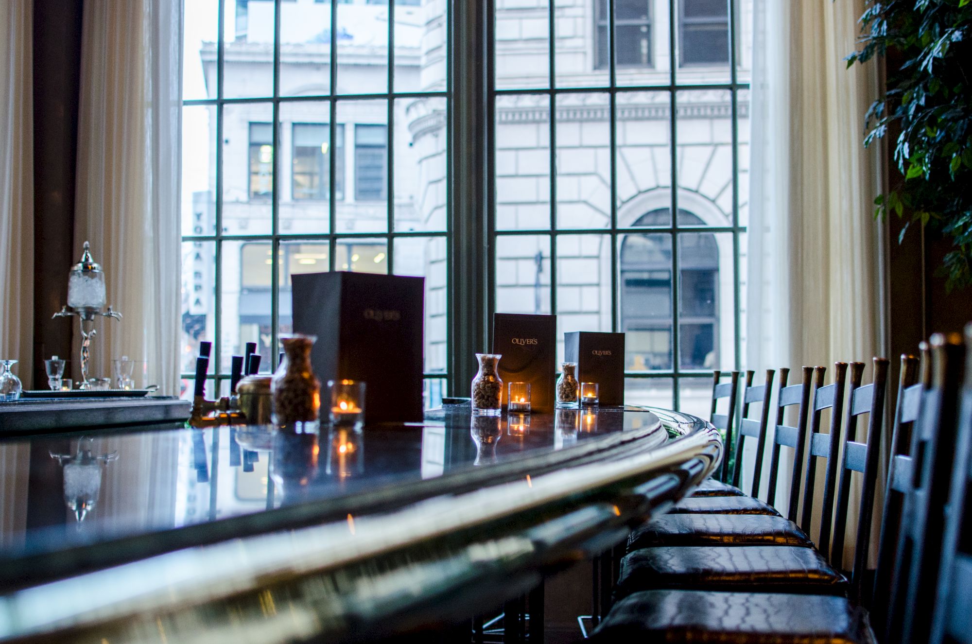 A long, polished wooden bar with stools, candlelit table setup, and large windows overlooking a city street.