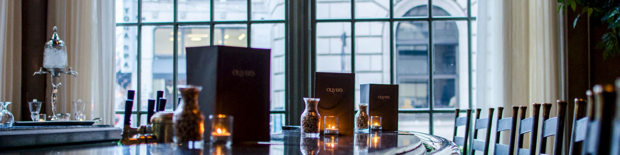 A long, polished wooden bar with stools, candlelit table setup, and large windows overlooking a city street.