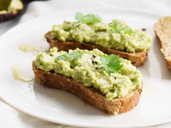 Two slices of toasted bread topped with mashed avocado, herbs, and a drizzle on a white plate, a simple avocado toast snack.
