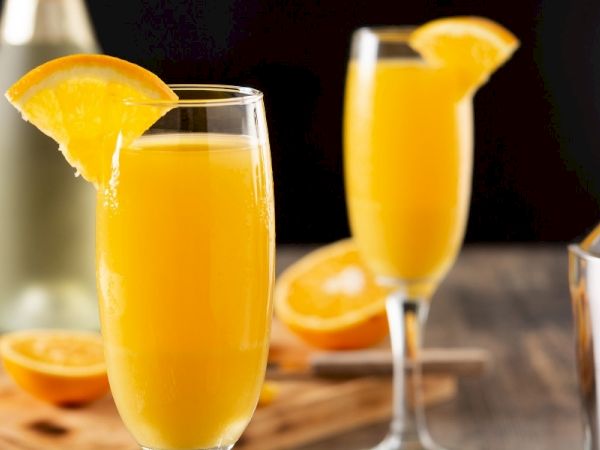 Two tall glasses of orange juice with orange slices on the rims, blurred bottles and halved oranges in the background.