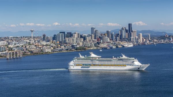 A large white cruise ship sails past a city skyline along a blue harbor, with tall buildings and distant mountains in the background.