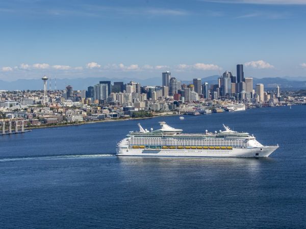 A large white cruise ship sails past a city skyline along a blue harbor, with tall buildings and distant mountains in the background.