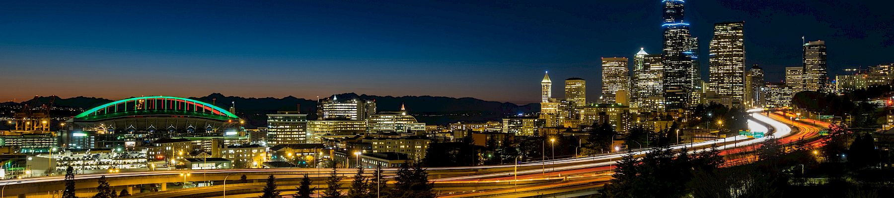 Nighttime cityscape with illuminated highways looping through a forested area, a distant skyline, and a dark blue-to-orange gradient sky.