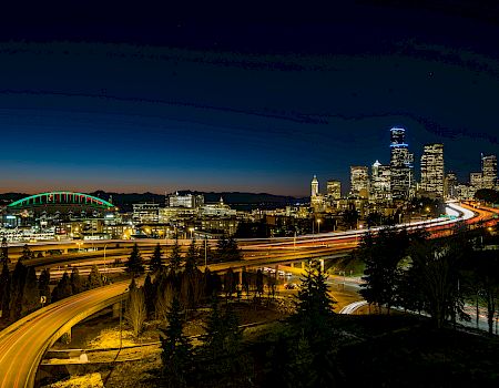 Nighttime cityscape with illuminated highways looping through a forested area, a distant skyline, and a dark blue-to-orange gradient sky.