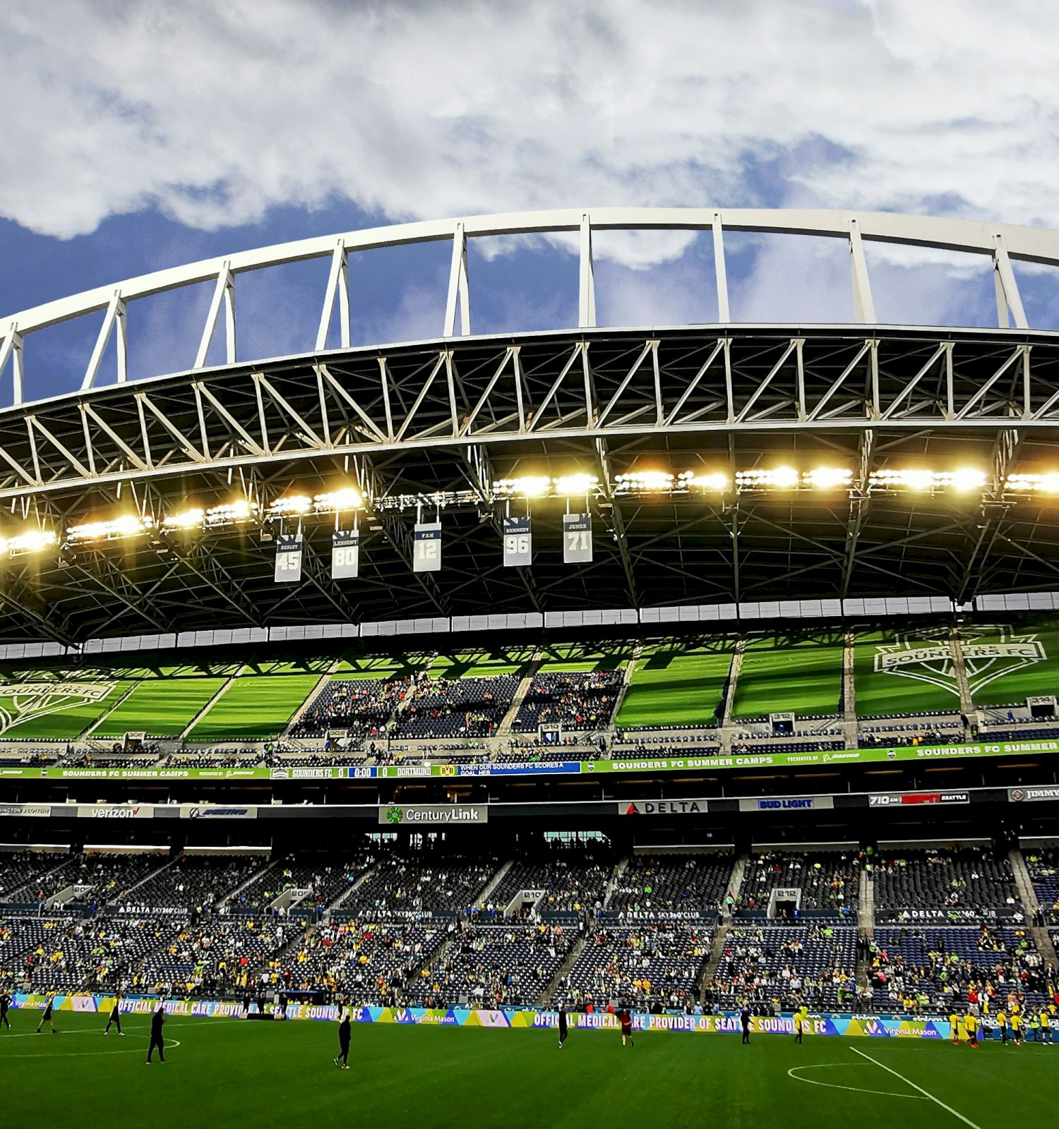 A large modern soccer stadium with green seating, a curved white roof, floodlights on, players on the field, and a partly cloudy sky, crowded stands.