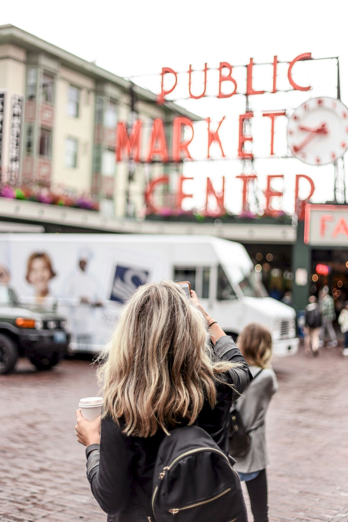 A woman with a backpack and drink in hand stands at a busy outdoor market, a large red &ldquo;Public Market Center&rdquo; sign overhead.