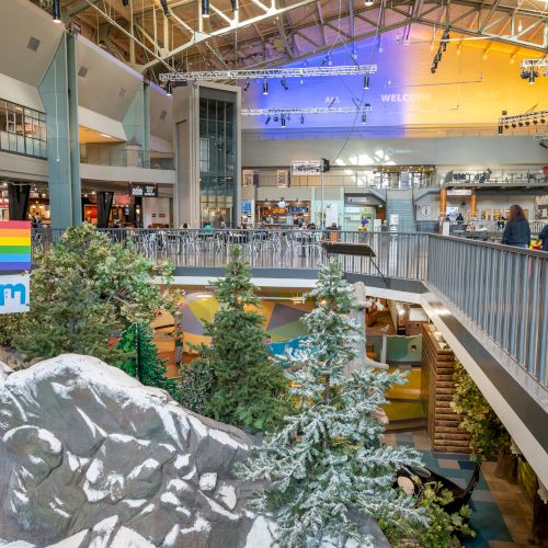 A bright, multi-level indoor space with a snowy mountain scene, pine trees, and people walking along upper and lower walkways at a mall or venue.