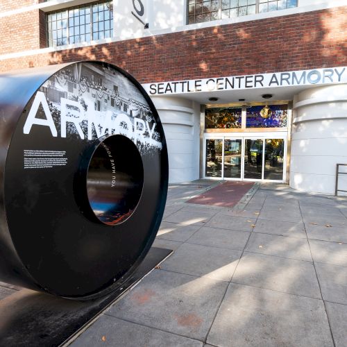A large black circular sculpture labeled &ldquo;ARMORY&rdquo; sits outside Seattle Center Armory, with the brick building and glass doors in the background.