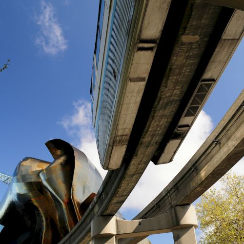 A modern elevated highway or rail structure seen from below, with steel beams and blue sky visible.