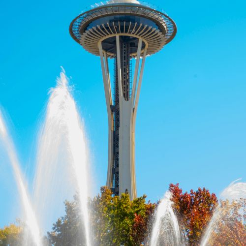 The Space Needle rises above a fountain in Seattle, with colorful trees below and a bright blue sky.