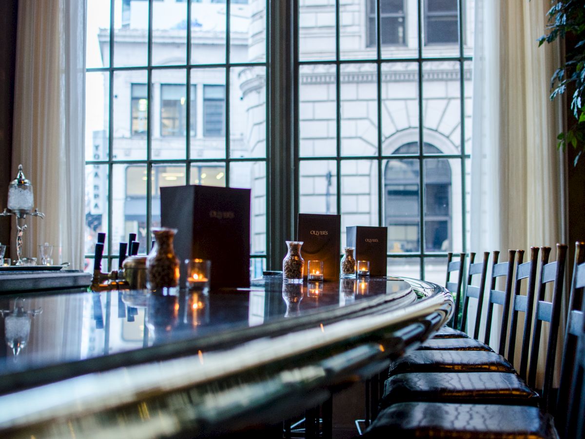 A long, curved bar with stools, candlelight, and a large window overlooking a city street, giving a cozy, upscale dining vibe.