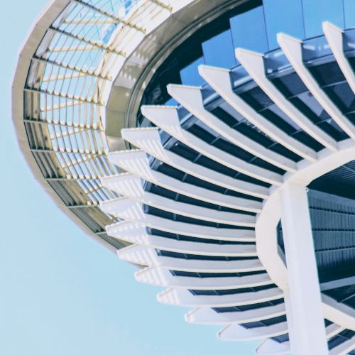 A futuristic white and blue tower with sweeping curved fins, glass canopy, set against a clear sky.