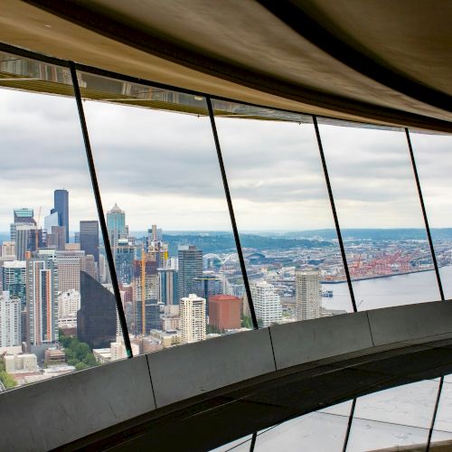 A city skyline seen through curved glass windows high above a river, with tall buildings and a cloudy sky, inside a modern observation deck.