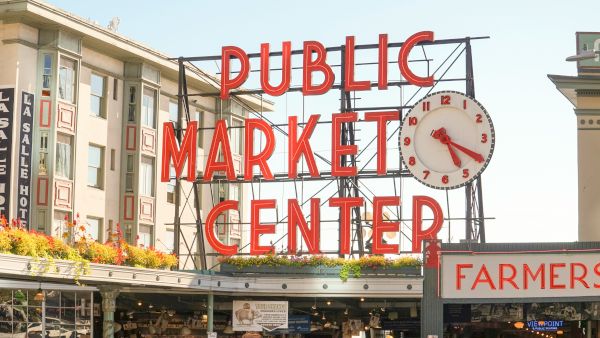 Public Market Center sign over a bustling market entrance with a large clock, Farmer&rsquo;s Market stalls, and city buildings in the background.